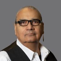 Indigenous man wearing black glasses, a white collared shirt, and a black vest.