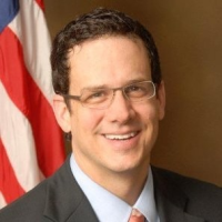 White man with short, dark brown hair and glasses standing in front of an American flag