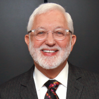 White man with white hair and beard smiling in a gray striped suit and black tie with colorful leaves